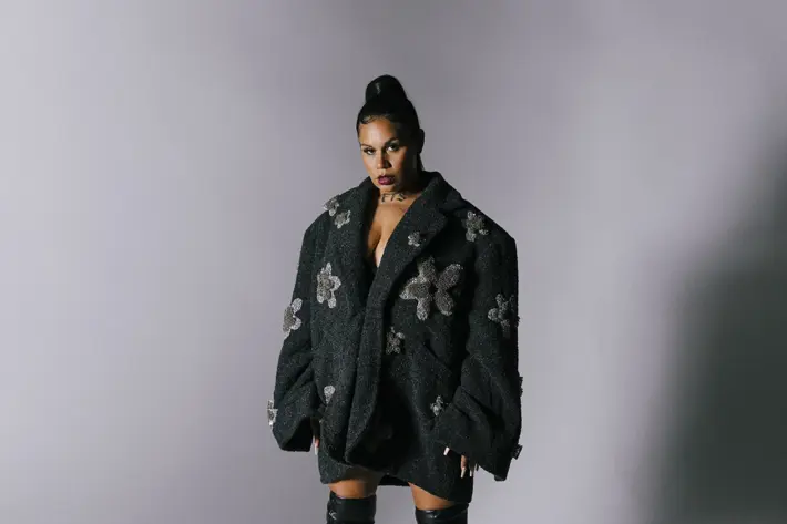 A person is standing against a plain grey backdrop wearing an oversized black textured coat decorated with large silver floral embellishments. The coat is styled as a dress, exposing the legs. The outfit is paired with thigh-high black leather stiletto boots. Soft lighting casts shadows on the floor and background, highlighting the dramatic and fashionable look.