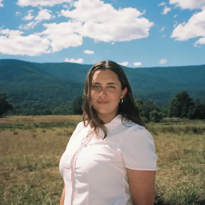 A person standing in an open grassy field with forested hills and mountains in the background under a bright blue sky scattered with white clouds. The person is wearing a short-sleeved white shirt and is positioned in the foreground, while the natural landscape stretches out behind them.