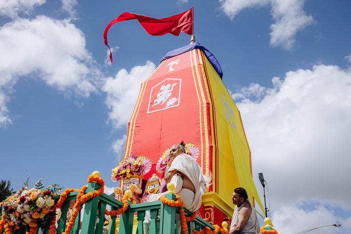 A vibrant outdoor parade features a large, decorated chariot with bright red and yellow fabric, adorned with colourful ornaments and traditional figures. The chariot is surrounded by a crowd of people walking along a street lined with palm trees and buildings. An Australian flag is visible near the chariot, and a billboard in the background reads “HAWAII FIVE-O.” The scene conveys a festive cultural celebration.